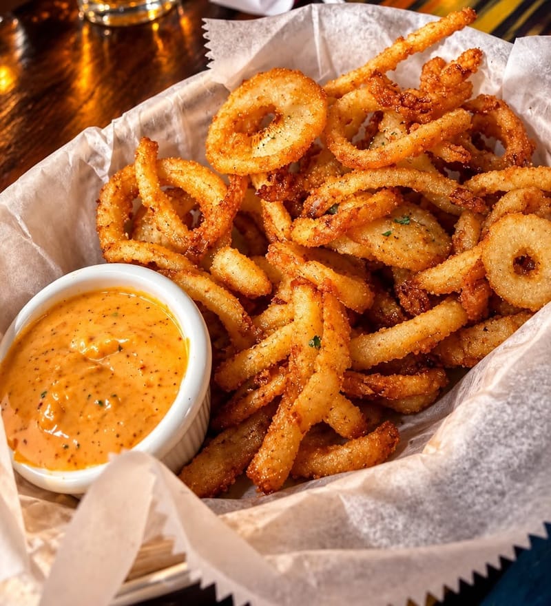 A basket of curly fries and onion rings with a cup of dipping sauce